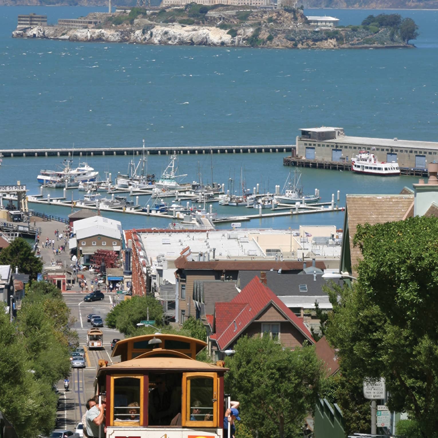 A trolly moving along a city street with water and island, and boats, in the distance.