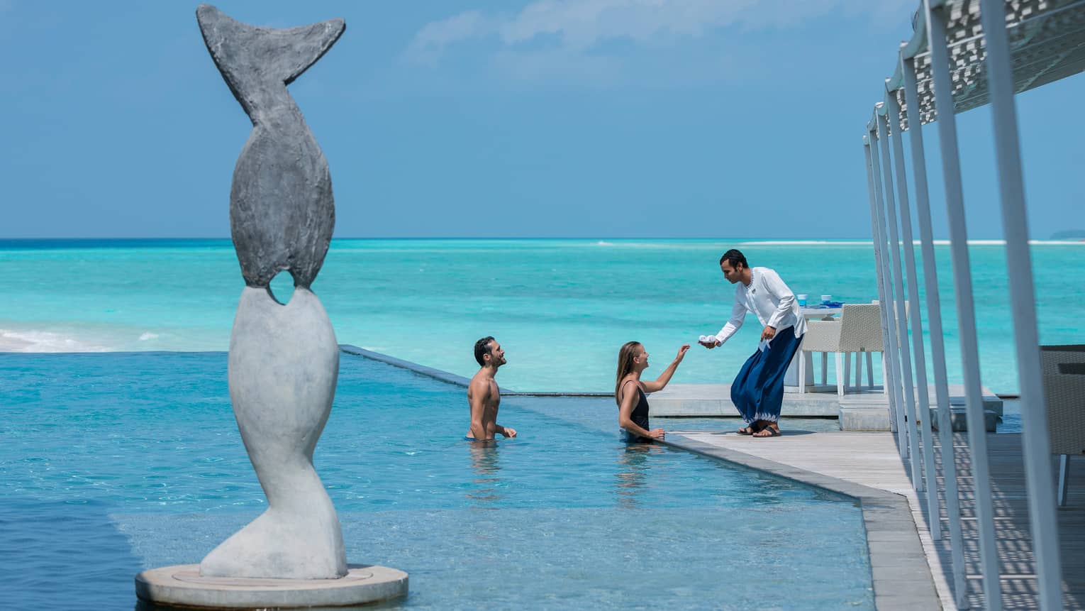 Server passes small towels to man, woman in swimming pool beyond tall stone statue of fish