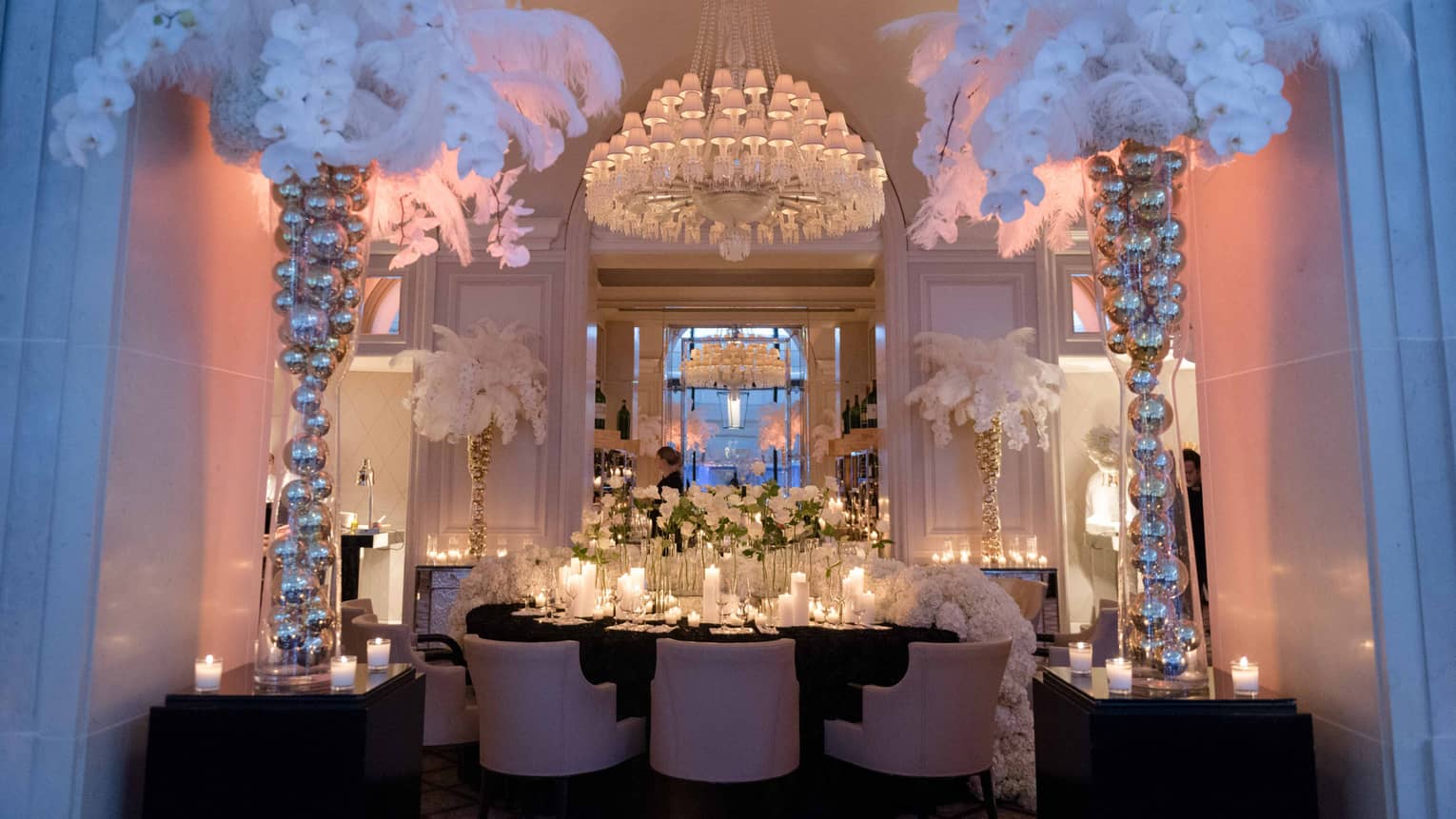Dining table under crystal chandelier, surrounded by tall vases filled with silver balls, faux white palms