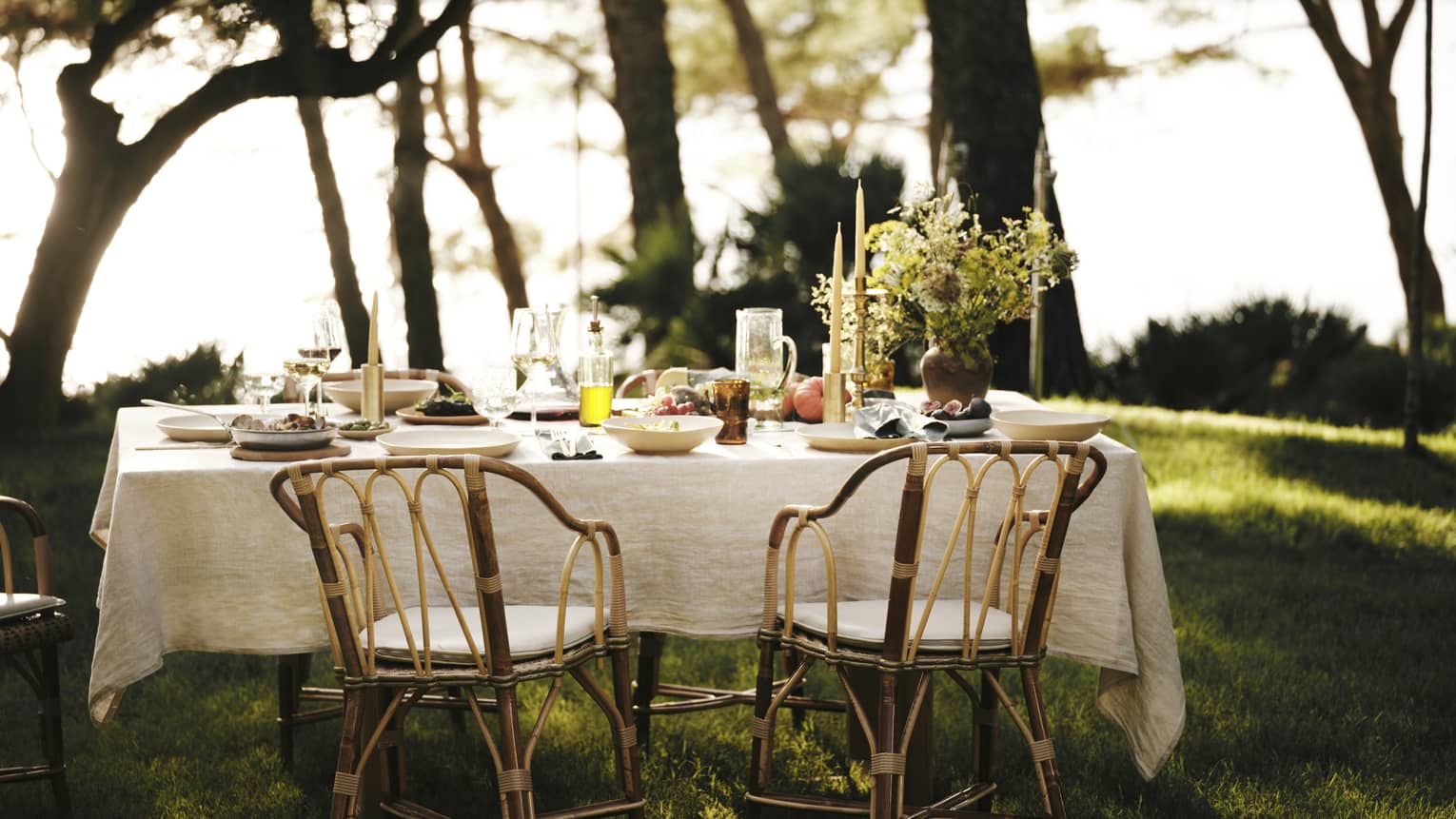 A linen tablecloth adorns an outdoor table set for five with wine, fresh seafood and fruit in an ocean-view forest setting.