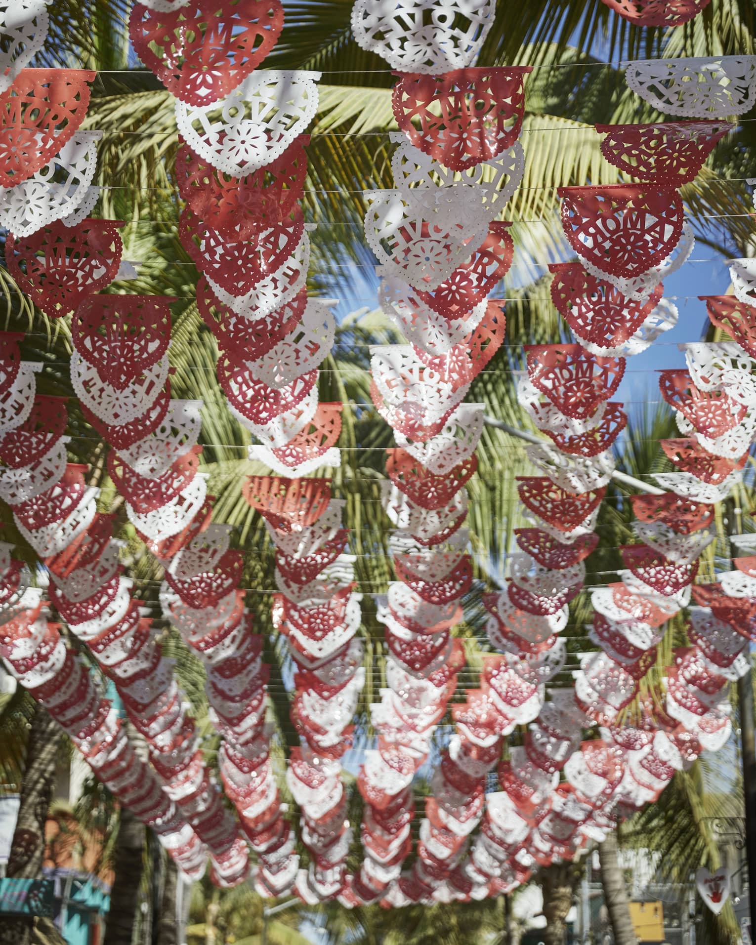 Looking up at rows of red and white Mexican Papel Picado banners hanging above, with palm trees and a sunny sky above them.