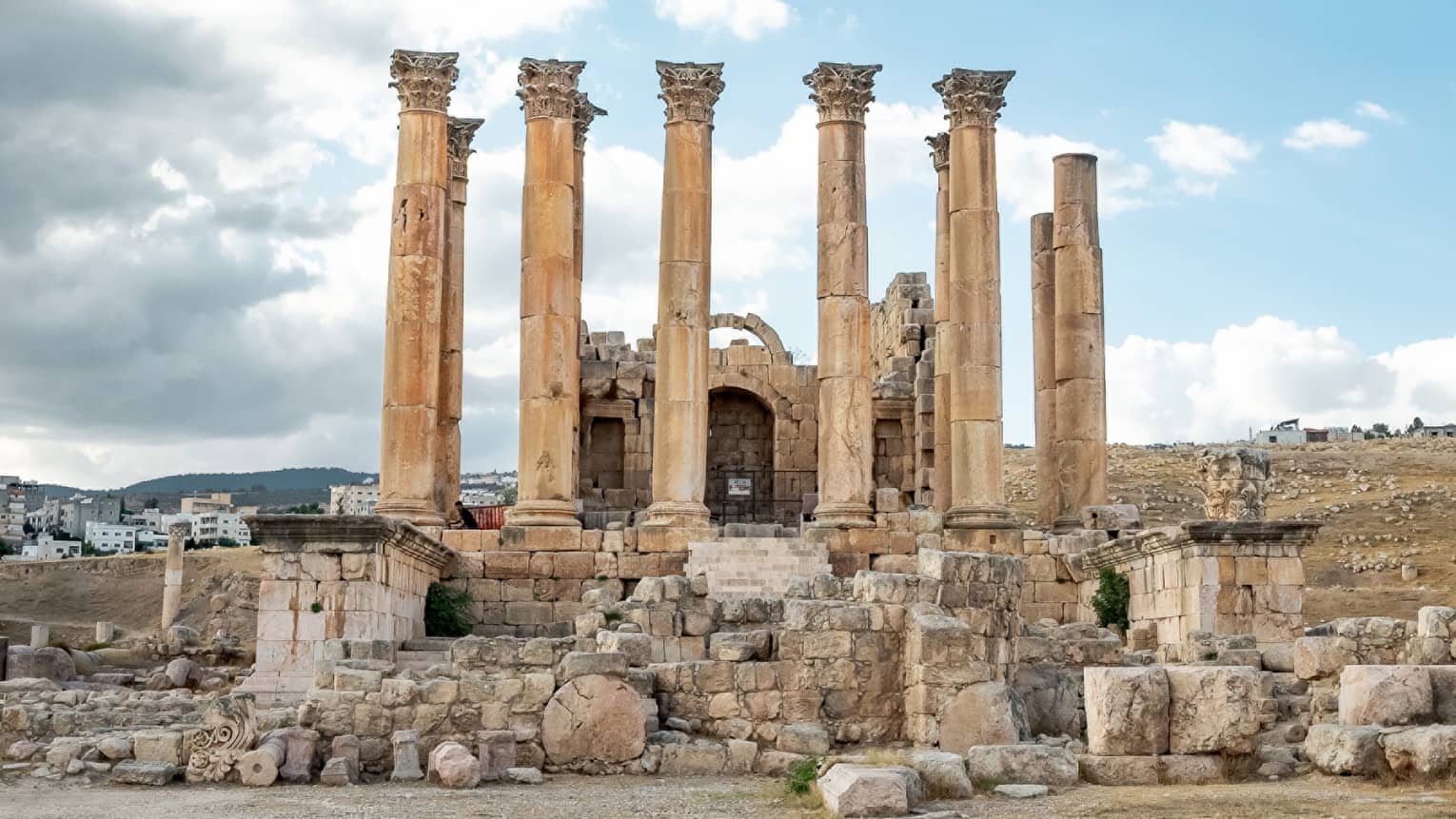 Amongst crumbling ruins, ten ancient Corinthian columns stretch skyward under a cluster of grey and white clouds.