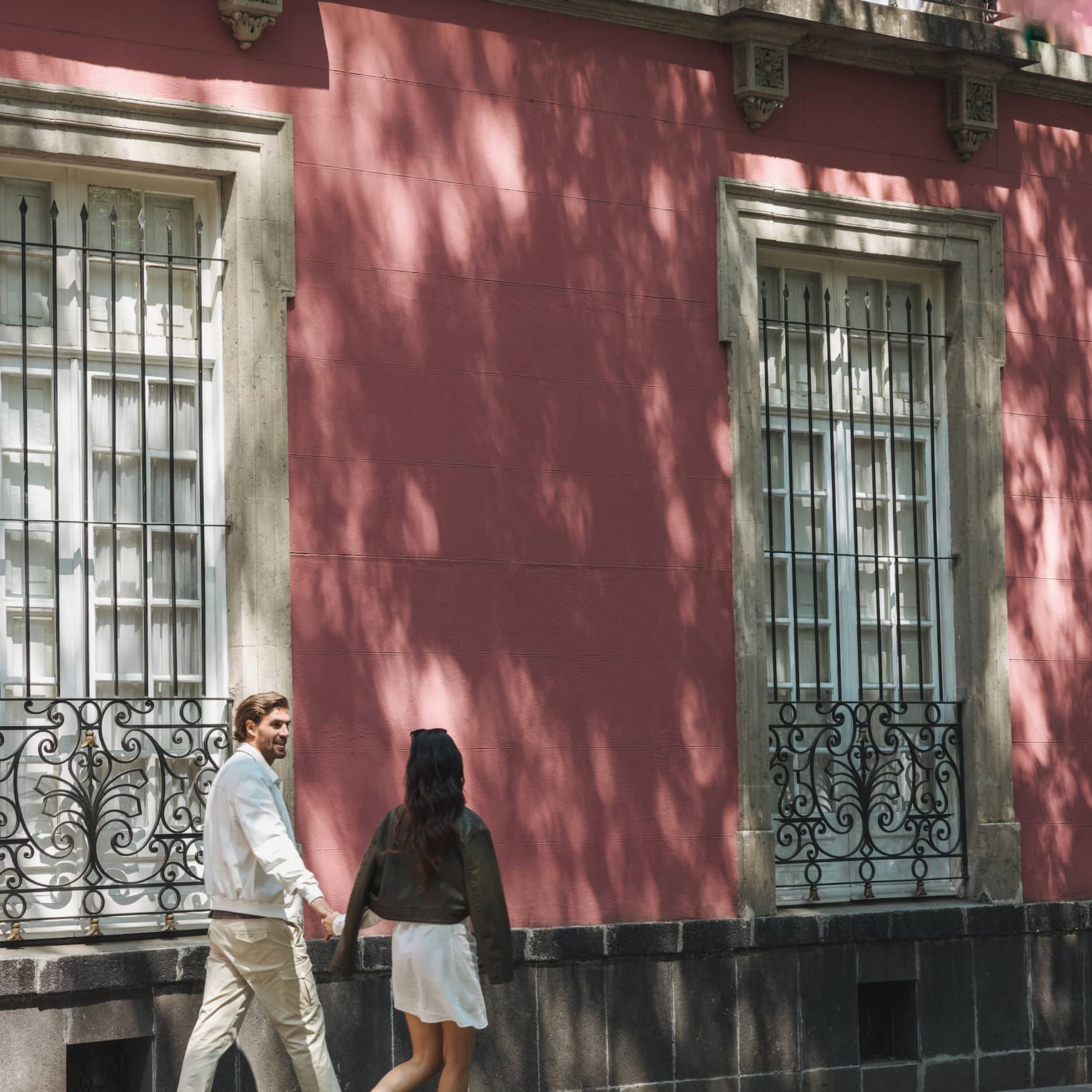 Couple strolls beside pastel pink bricked building featuring tall, paned windows adorned with ornate wrought iron railings.