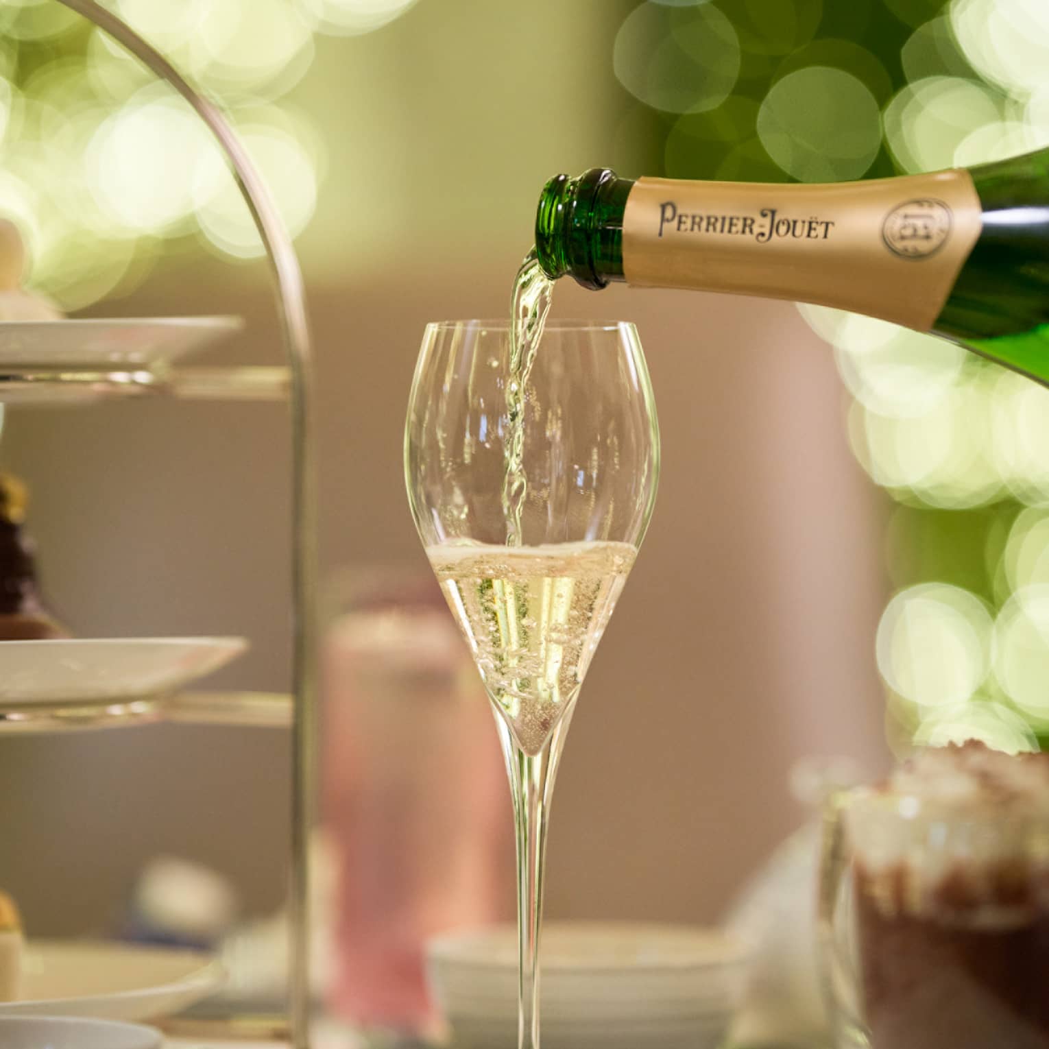 Champagne is poured from a bottle of Perrier Jouet into a glass next to a tiered tray of treats and a frothy cup of espresso.