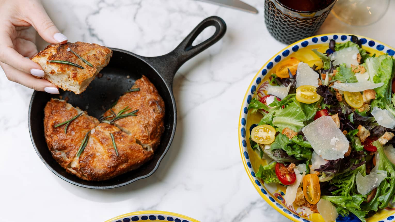 Amid colourful dishes, cutlery and glassware, a hand takes focaccia from an iron skillet, another takes a forkful of ravioli.