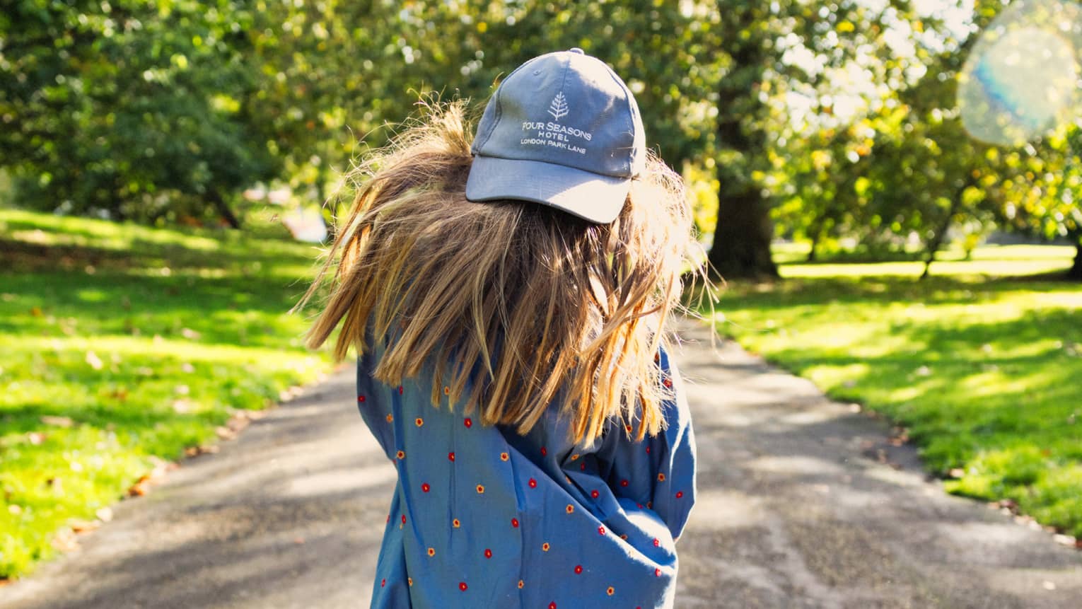Rear view of long-haired child on a sunny path flanked by green grass and trees, wearing backwards London Park Lane ball cap.