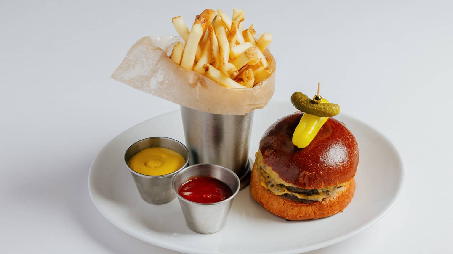 Burger topped with a yellow pepper and gherkin pickle on a toothpick plated next to a silver cup filled with French fries and small tins of ketchup and mustard