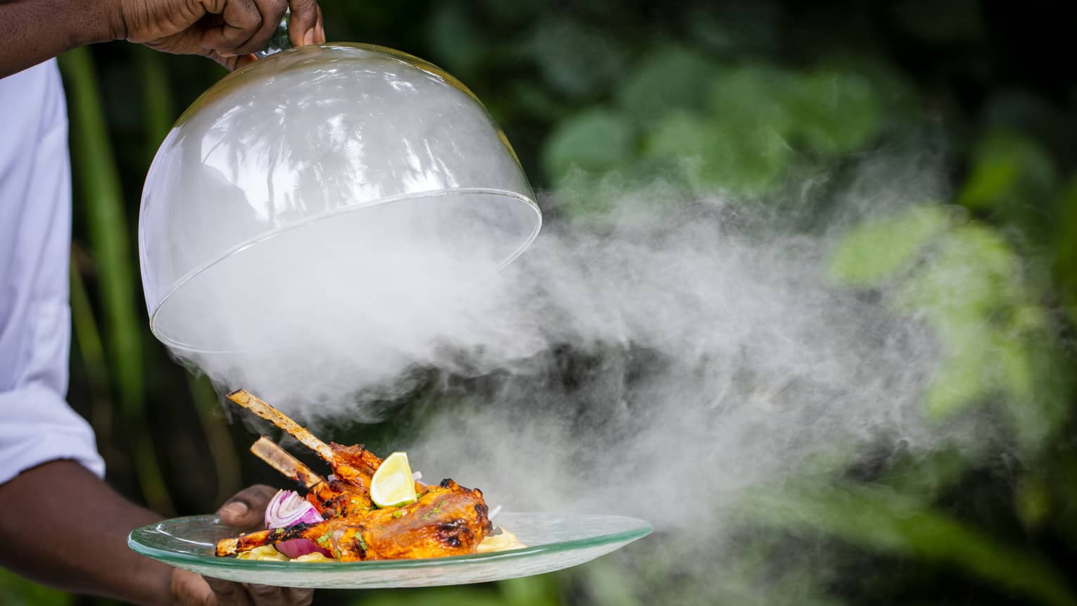 A Four Seasons dining staff taking the lid off of a dinner tray – steam is pouring out over a cooked meat dish on skewers
