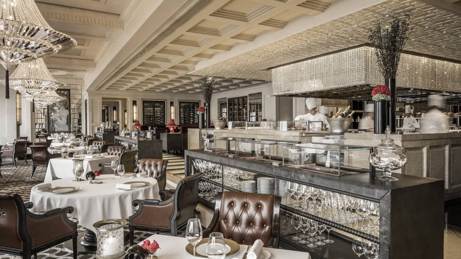 An elegant French restaurant interior with polished wooden floors, white tablecloth-covered tables, and plush leather chairs. The room features chandeliers. 