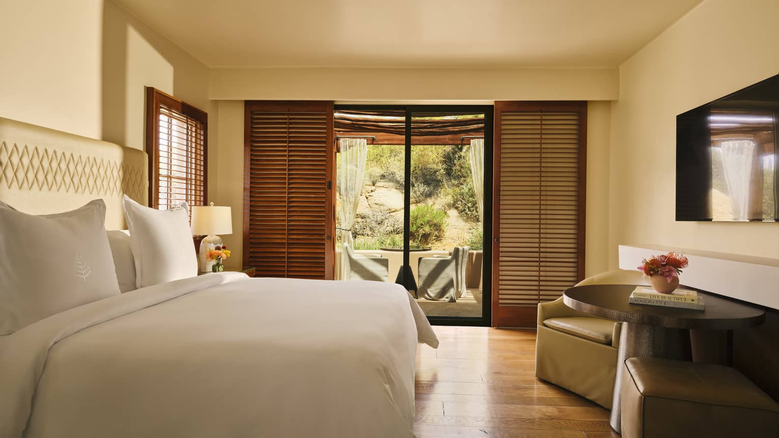 Bedroom with a king bed, two arm chairs and small table, TV, wooden floors and walk-out terrace with shuttered doors
