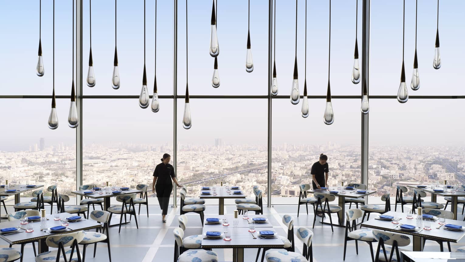 Large glass raindrop-shaped lights hang from soaring ceiling in sunny dining room, servers set tables