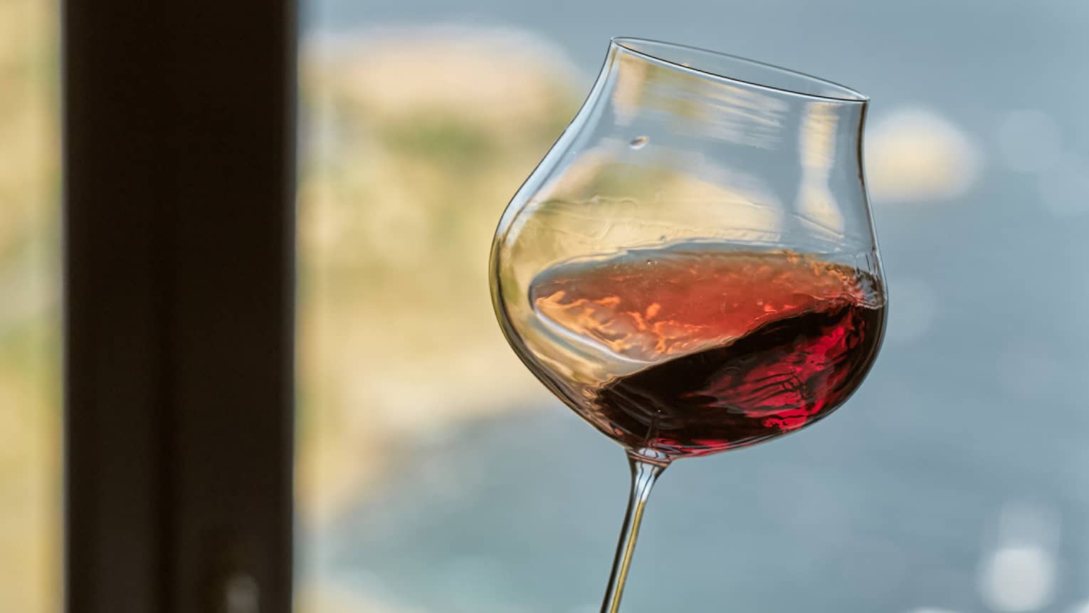 In front of a window, a hand holds a stemmed glass at the base and swirls red wine. Blurred seashore in the background.