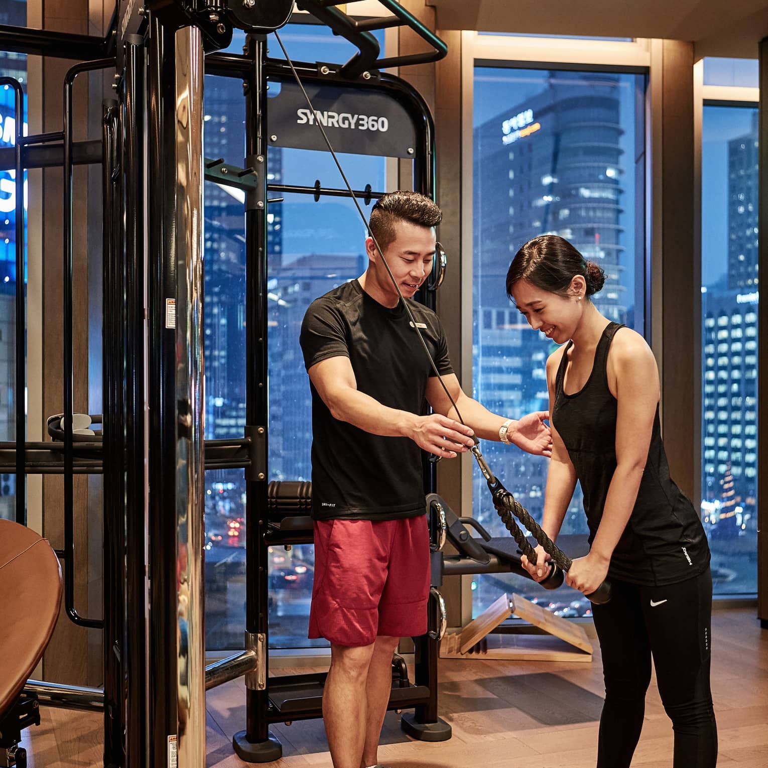 With skyscrapers out the window beyond, a trainer guides a guest as she performs a triceps pushdown exercise on a gym machine.