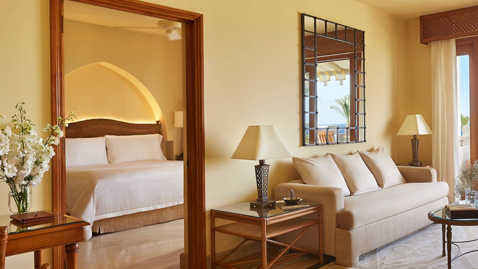Hotel room seating area with white sofa, armchair, area rug, open balcony door, door to bedroom