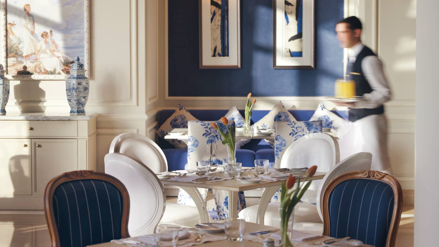Kala Restaurant server in uniform vest with tray of orange juice walks past white-and-blue dining chairs