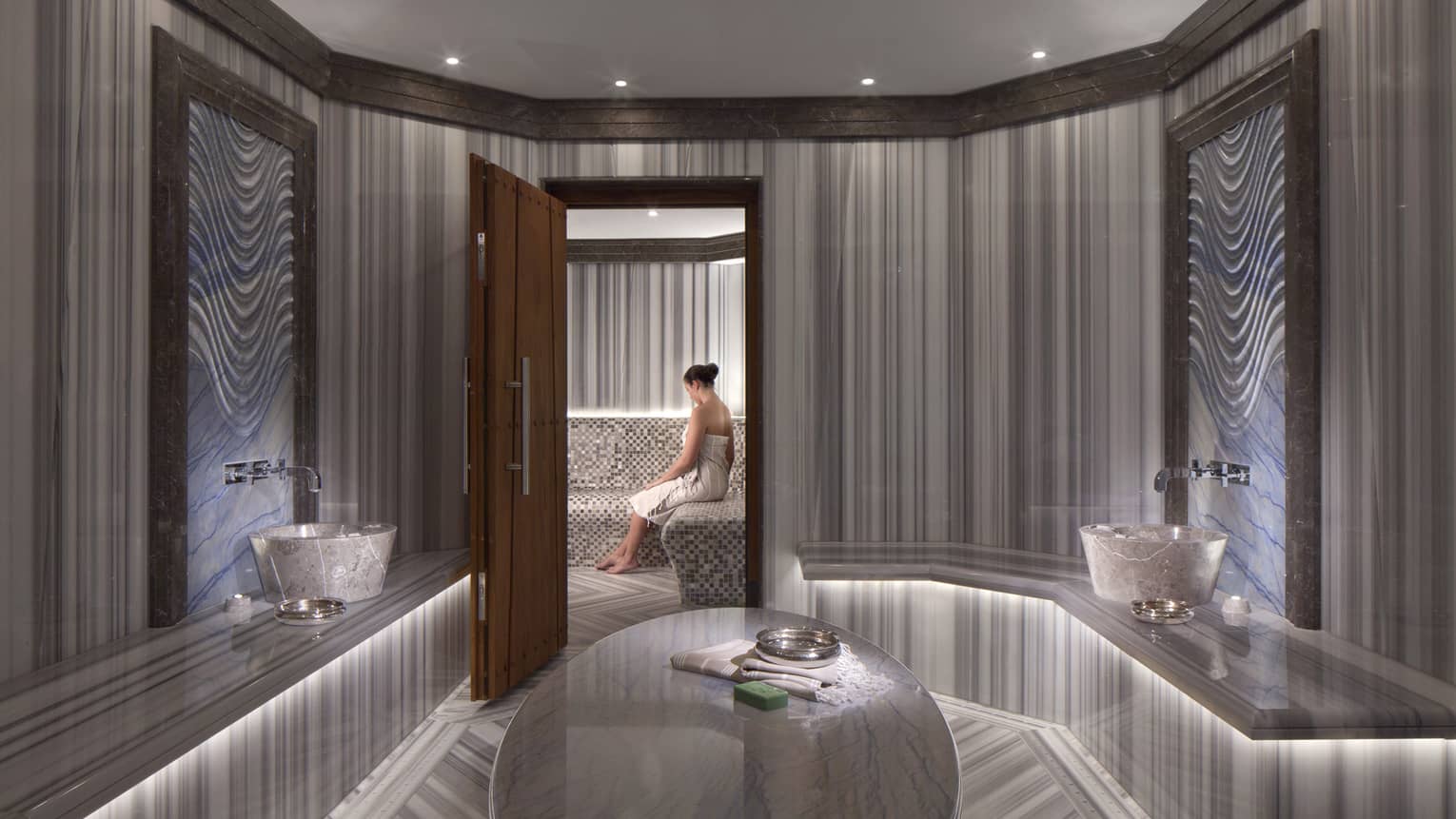 Elegant marble spa room with open wood door where woman wearing towel sits on bench 