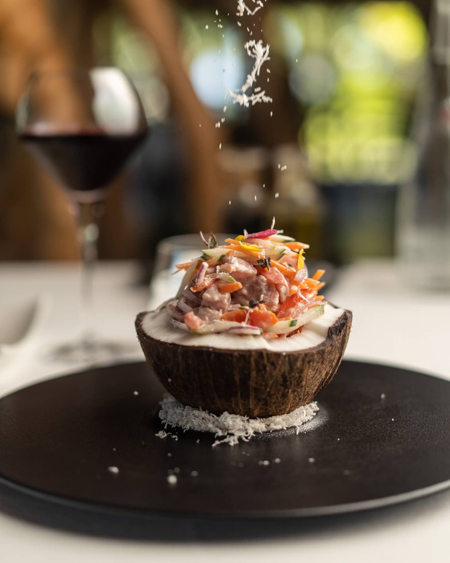Close-up of an open coconut filled with fish and thinly sliced vegetables, atop a bed of coconut flakes on a round platter.