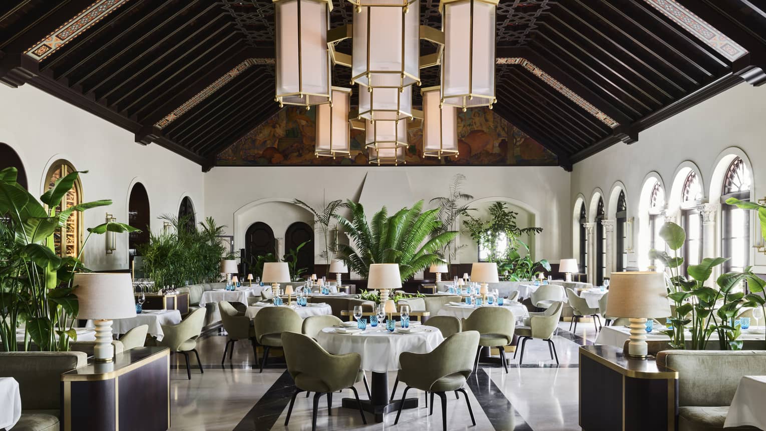 A sophisticated dining room with high, dark wood-beamed ceilings, large hanging lantern chandeliers, green upholstered chairs, white tablecloths and indoor plants.