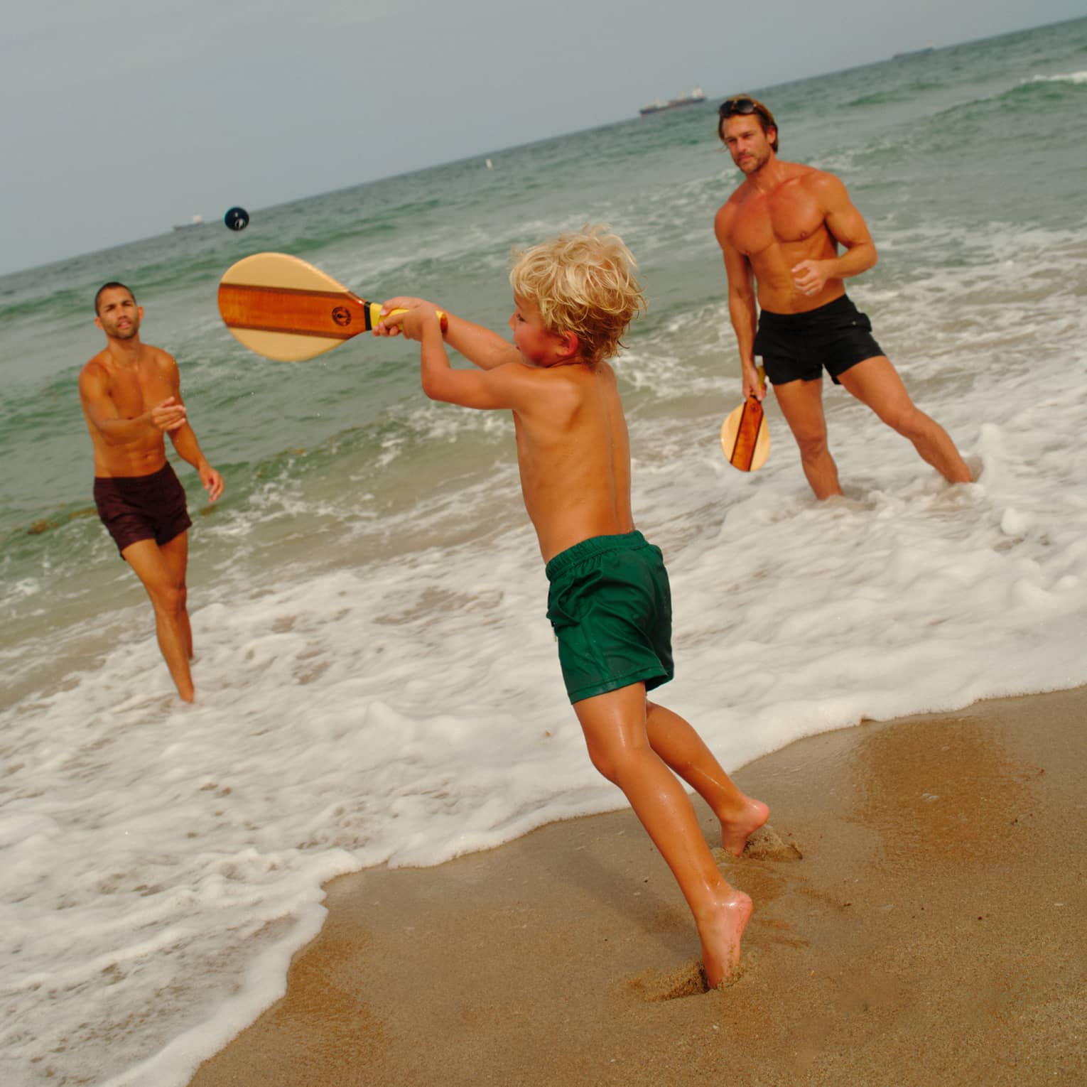 A young shild plays paddle ball in the surf with two adults