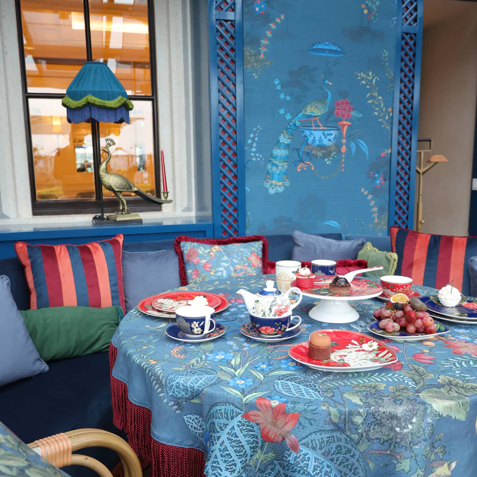 A peacock inspired décor of blues, greens and reds around a table set up for afternoon tea with plates of fruit and pastries.
