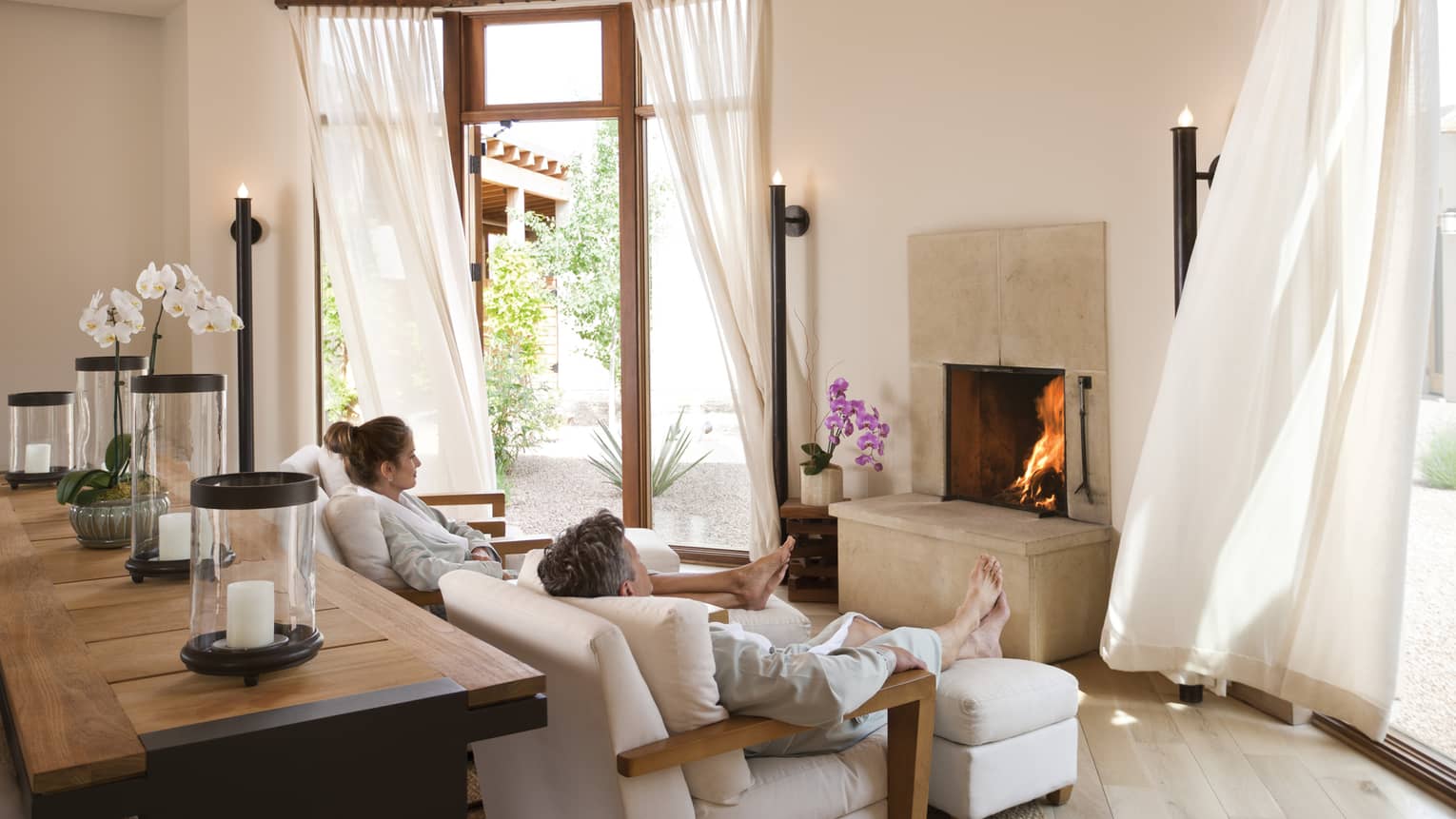 Man, woman in white bathrobes relax on spa armchairs by fireplace, white curtains