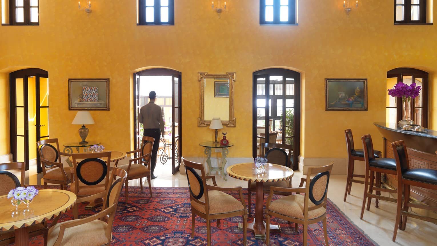 Man walks out of open French doors from bright lounge with yellow walls, dining tables, bar