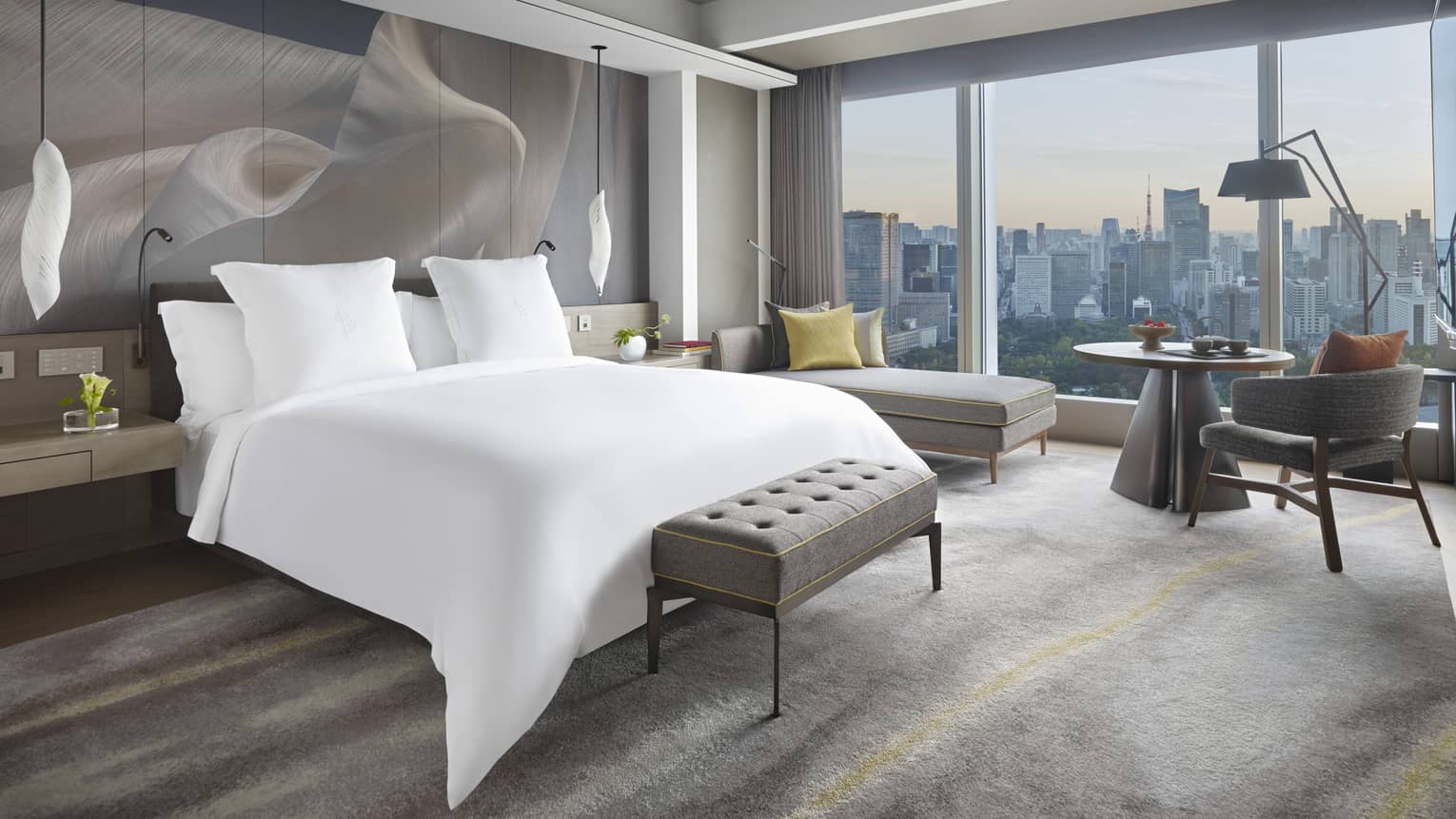Guest room with king bed, tufted bench, chaise lounge, table with armchair, pendant lights and large window offering city views