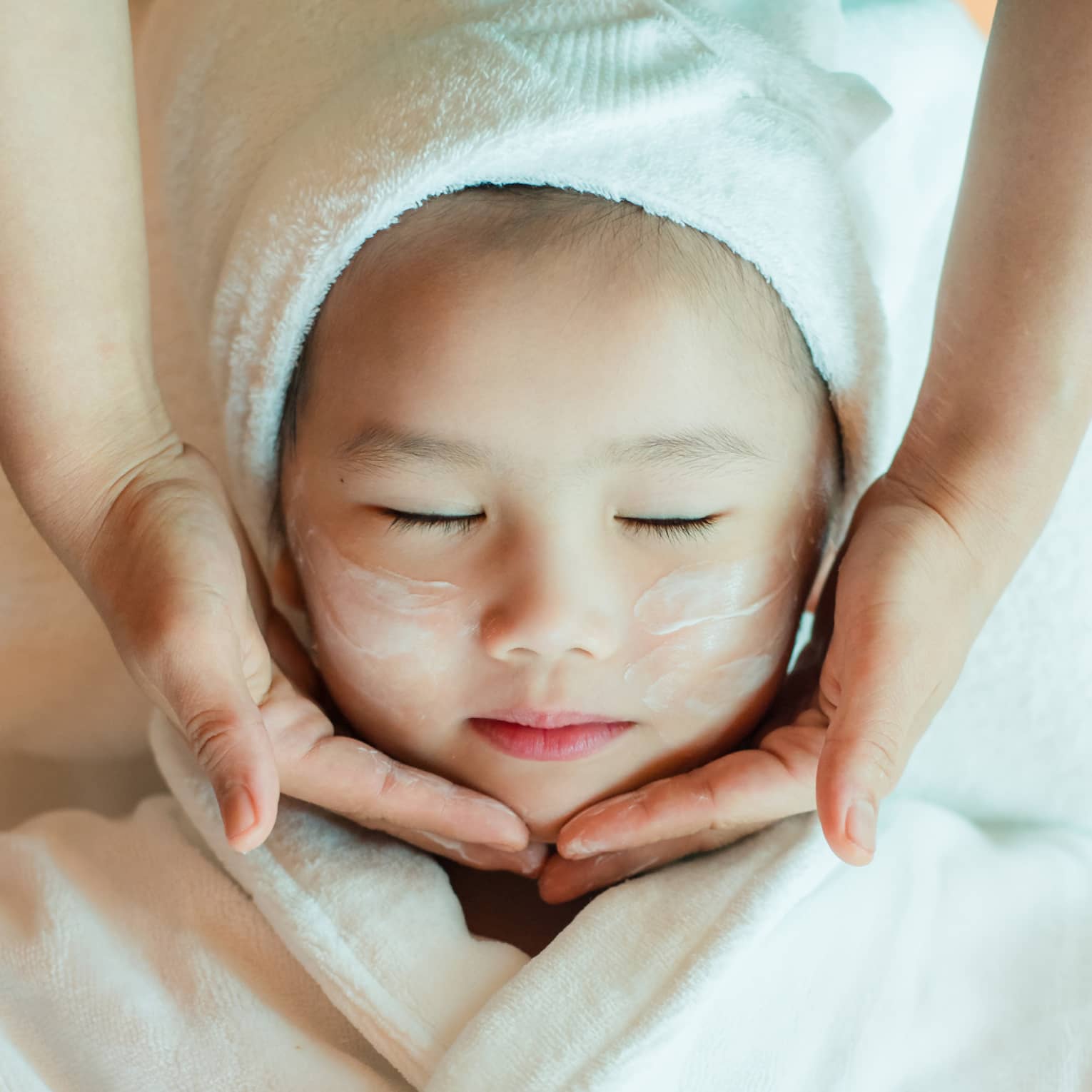 A close-up of a relaxed child wrapped in a towel and robe and enjoying a masseuse massaging a cream into their face.