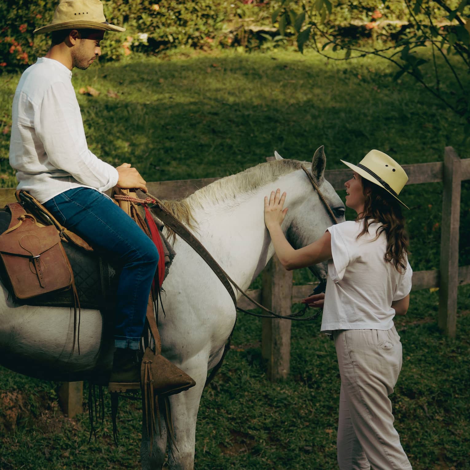Person wearing a white long-sleeve shirt and jeans sits atop a white horse as another person wearing a white short-sleeve top and cream pants stands on the ground with one hand on the reins and another on the horse