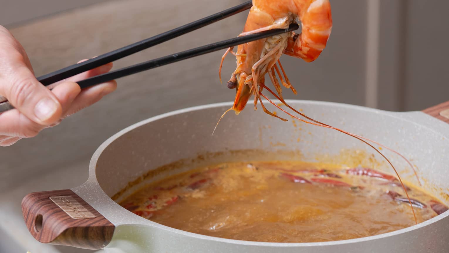 Chopsticks lift a piece of shrimp from a simmering pot on a hotplate, next to a small bowl of mushrooms and lettuce.