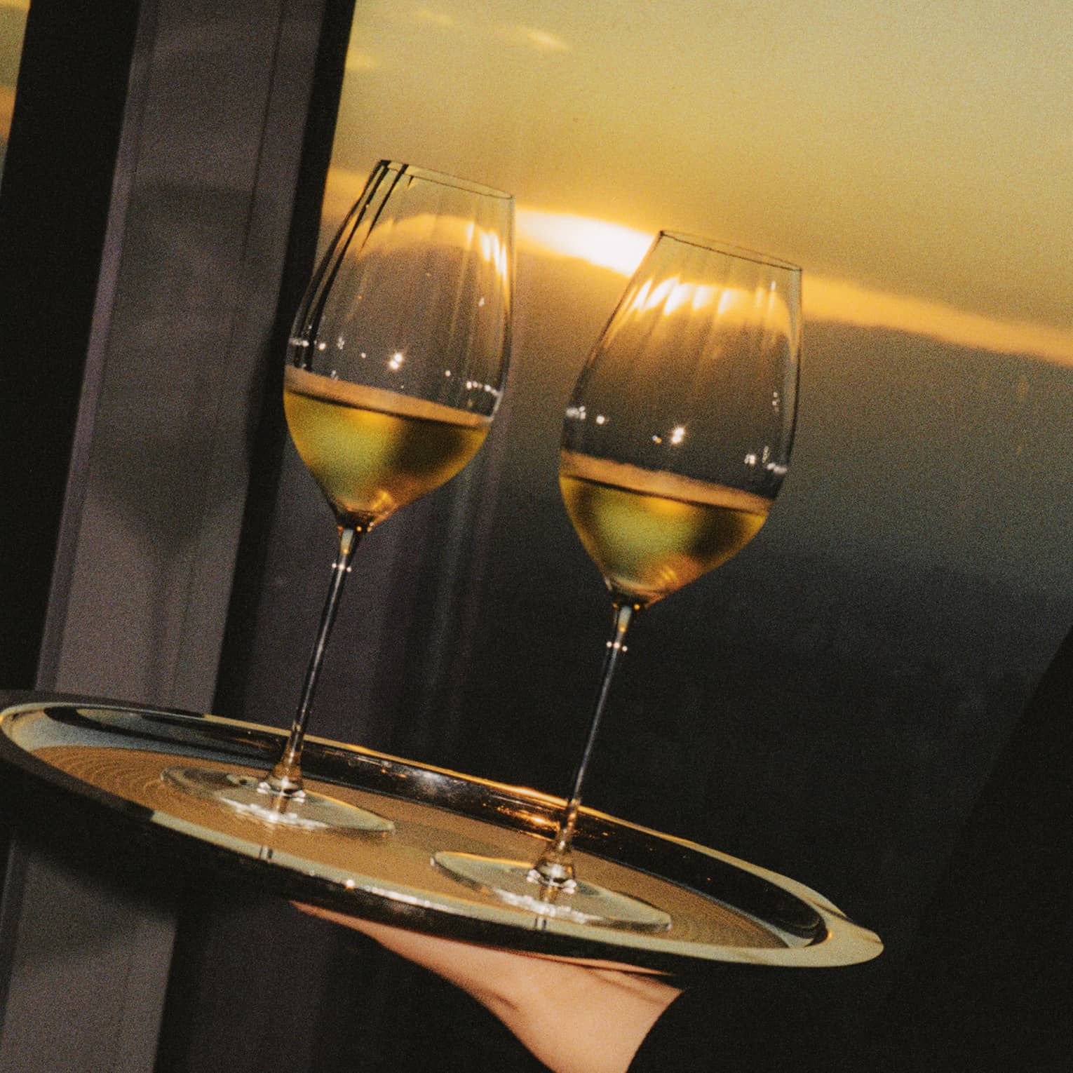 Two glasses of wine being served on a hand-held tray with the sun shining in the background