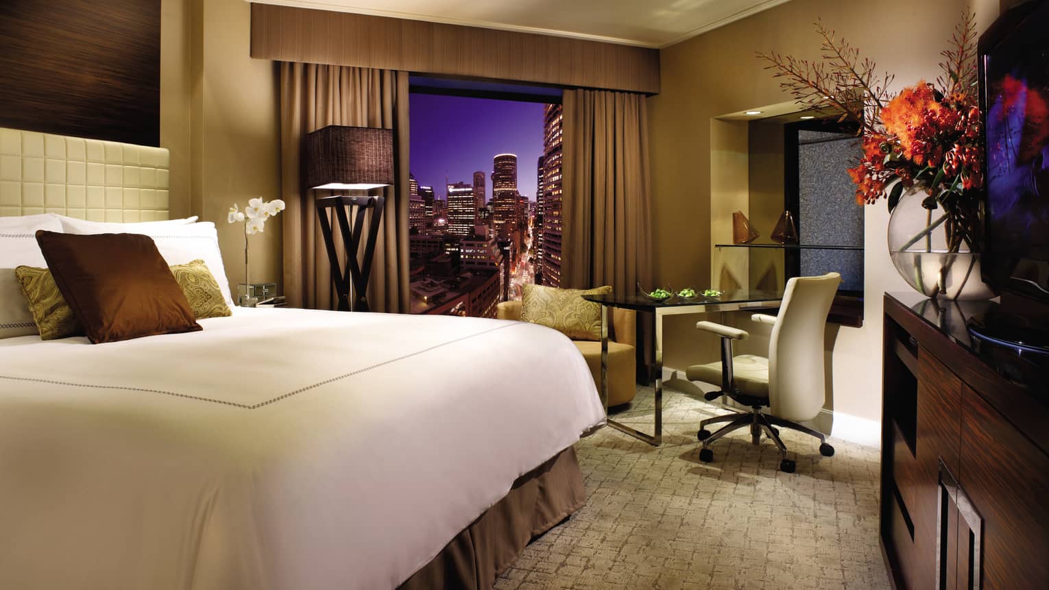 Deluxe City Room at night, bed with brown silk pillows, large glass vase with flowers, window with city view