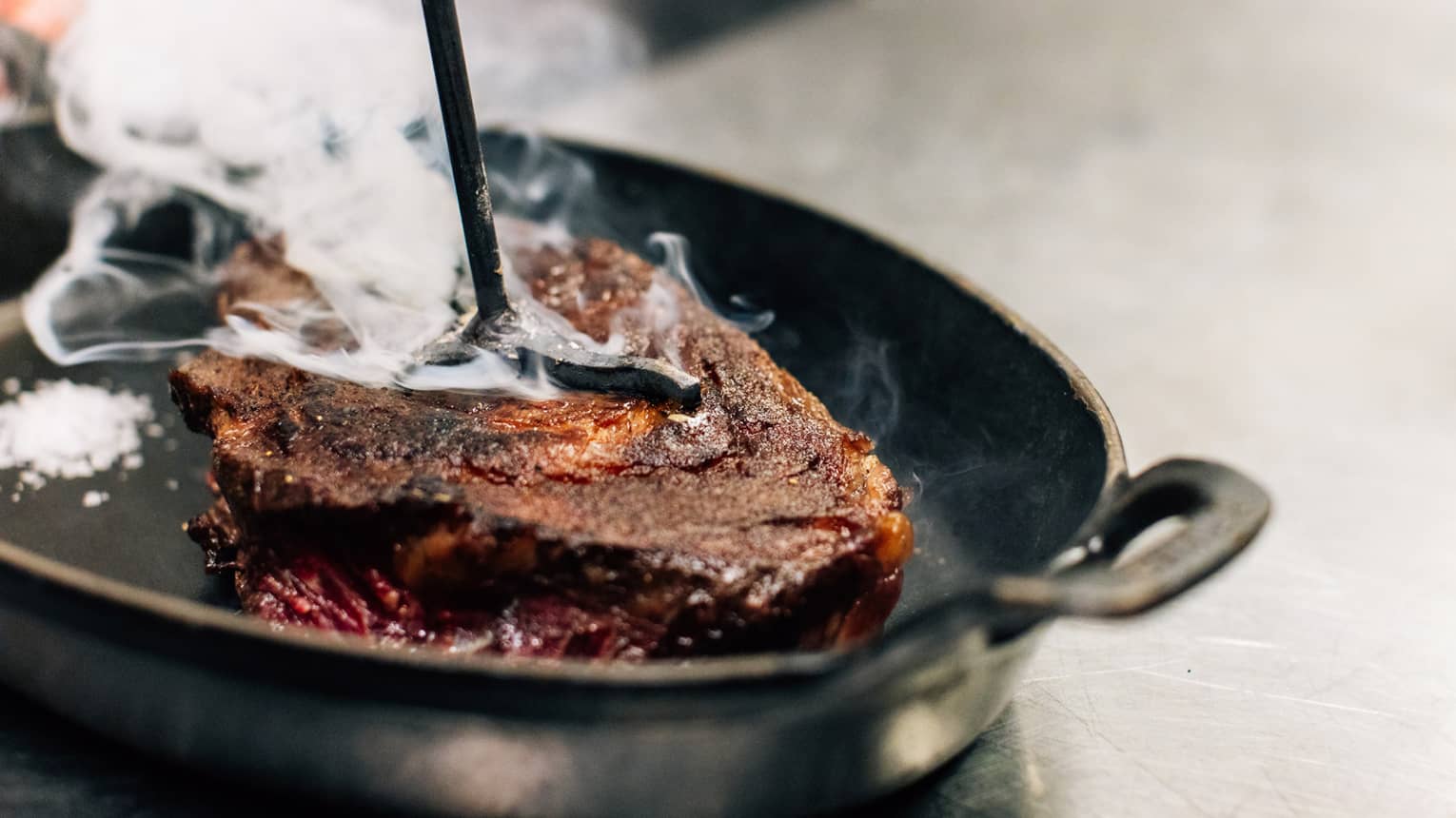 A close up of a Longhorn-Branded Steak steaming in a cast iron skillet on a silver, metal industrial counter