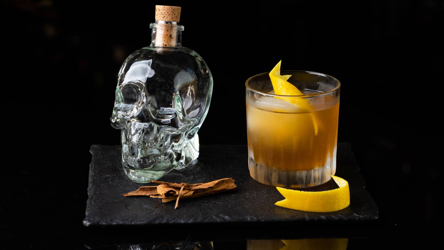 A skull glass container with a glass of orange cocktail next to it.