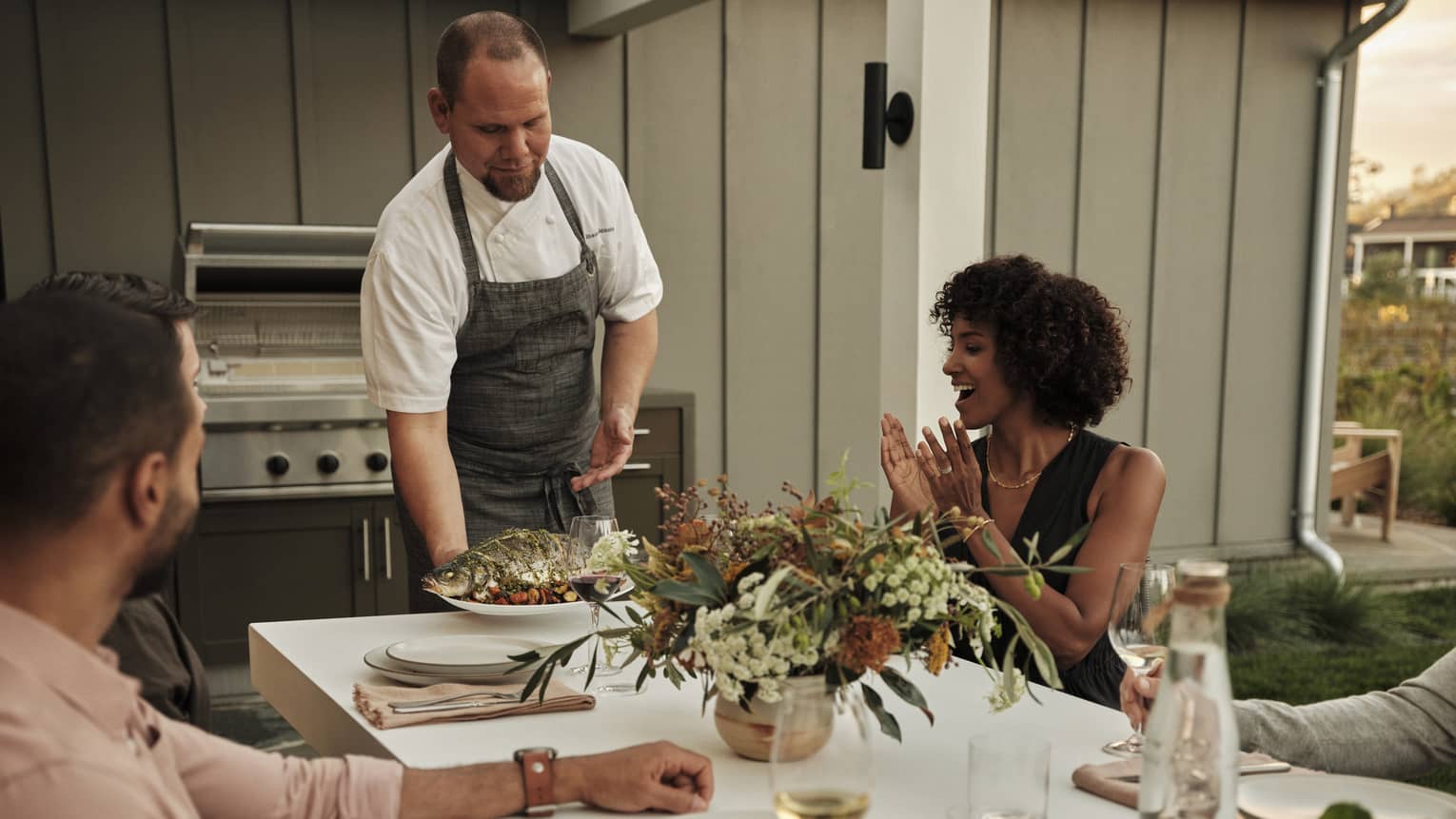 A guest smiles and claps as a chef sets down a plate of grilled fish at an outdoor table decorated with earth-toned flowers.