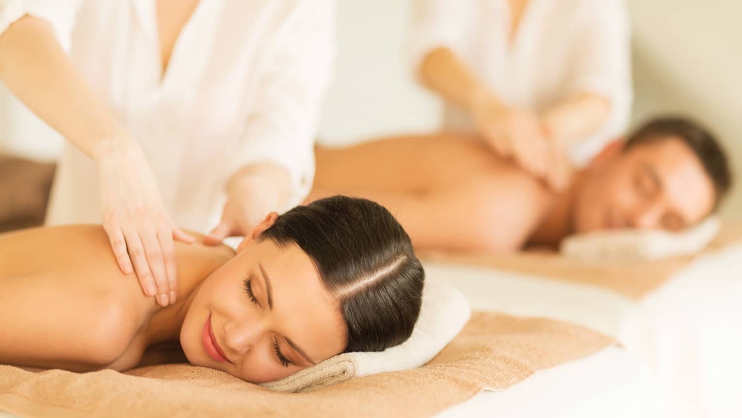 Woman and man lie side-by-side on massage tables as spa staff massage their bare shoulders