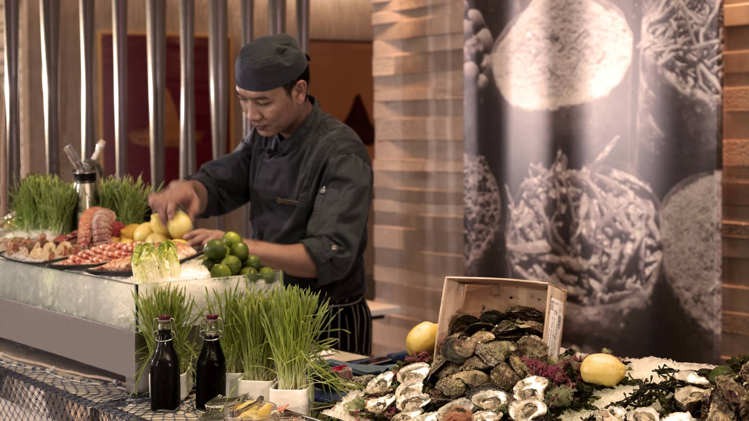 Man in black cook's uniform stands behind buffet with platters of fresh raw seafood on ice, citrus fruit