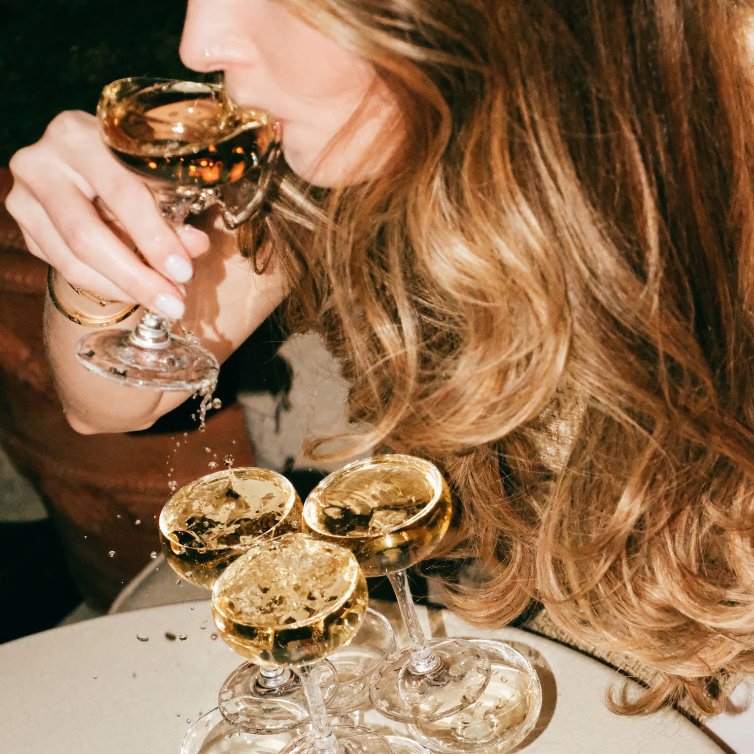 Person with long sandy blonde hair sips champagne from a coupe glass taken from the top of a tower of stacked coup glasses all filled with champagne