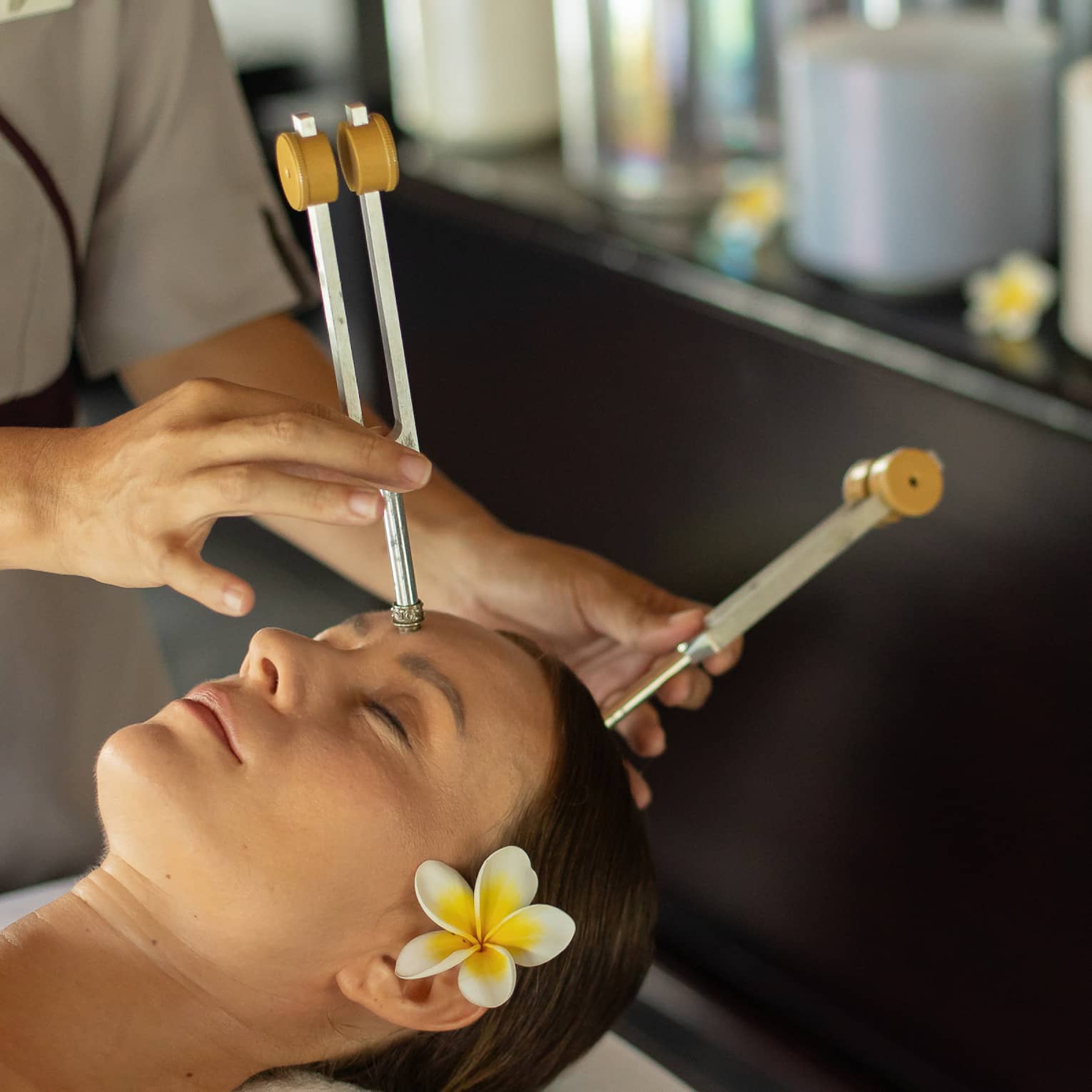 A practitioner delicately applies ohm tuning forks to the forehead and crown of a guest resting peacefully on a massage bed.