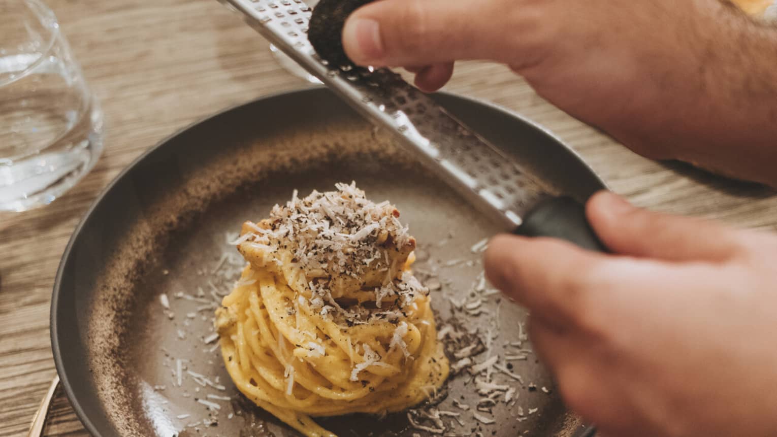 A person grates nutmeg over a creamy pasta dish topped with cheese