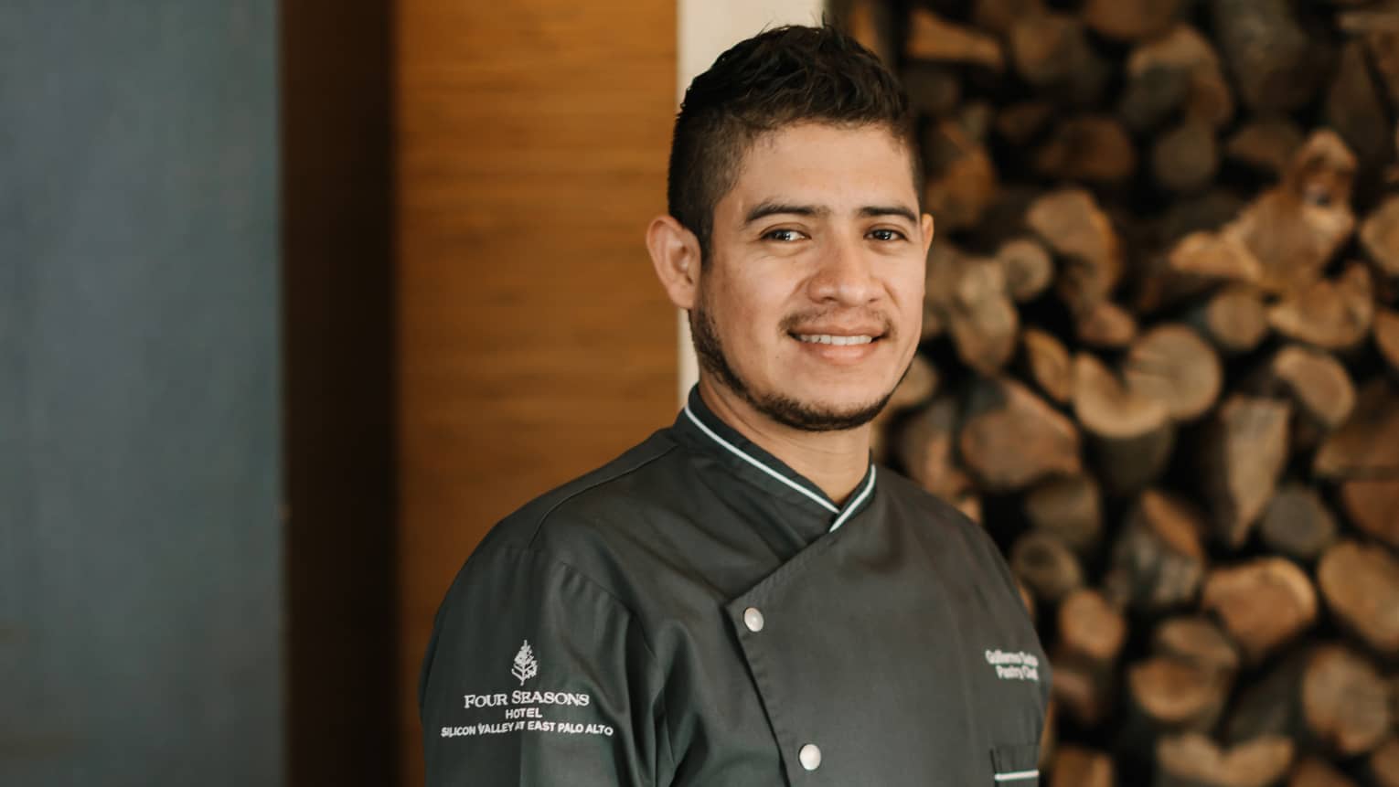 A chef in grey chef clothes standing in front of a pile of cut fire wood.