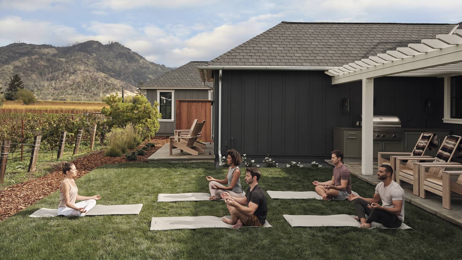 Group of people practicing yoga on mats in a grassy villa backyard near vineyards, with mountain views, wooden chairs and an outdoor grill area under a pergola