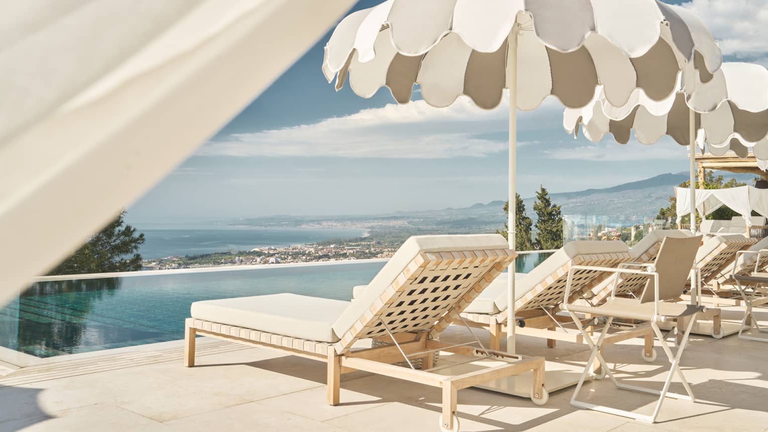 An infinity-edge pool with a bit of cabana draping in the foreground. Lounge chairs and umbrellas line one side of the pool, and land and ocean views are in the background.