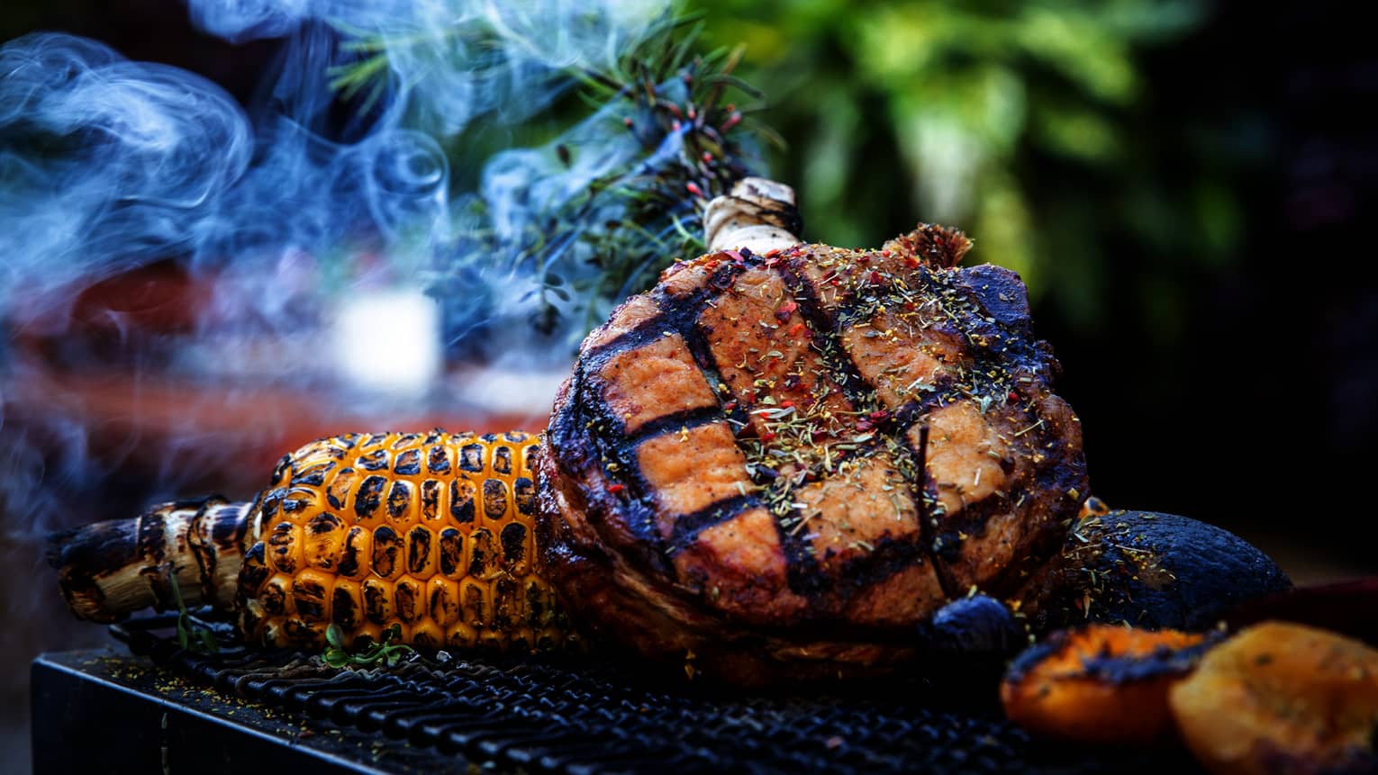 A grill with meat, corn and potatoes cooking on it outside.