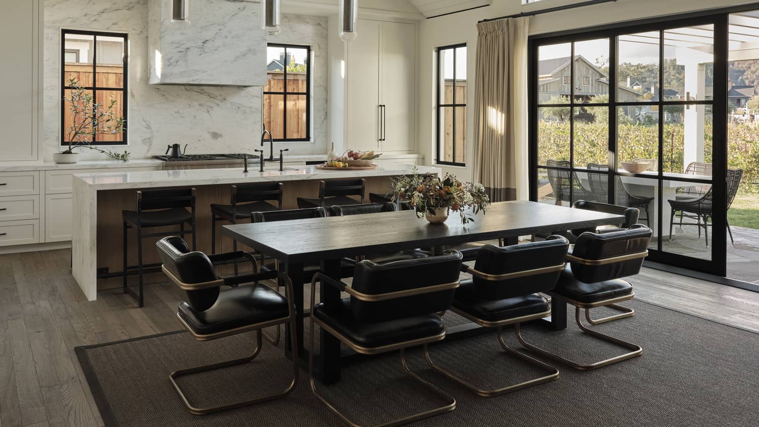 Open kitchen and dining area with marble island, dark wood table, black leather chairs, pendant lights, windows and sliding doors leading to outdoor patio