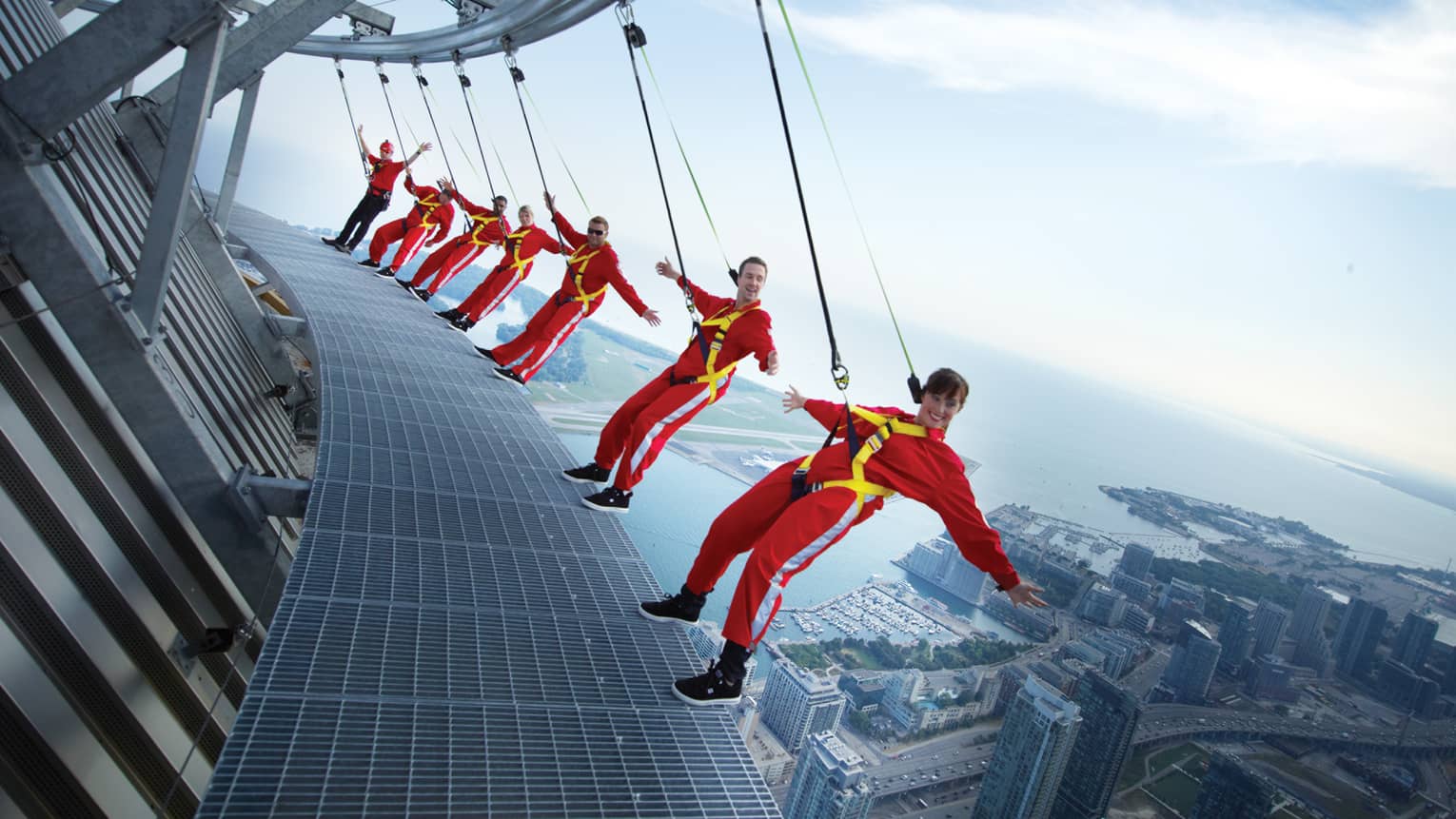 People wearing red jumpsuits, harnesses lean back over edge of CN Tower high above city