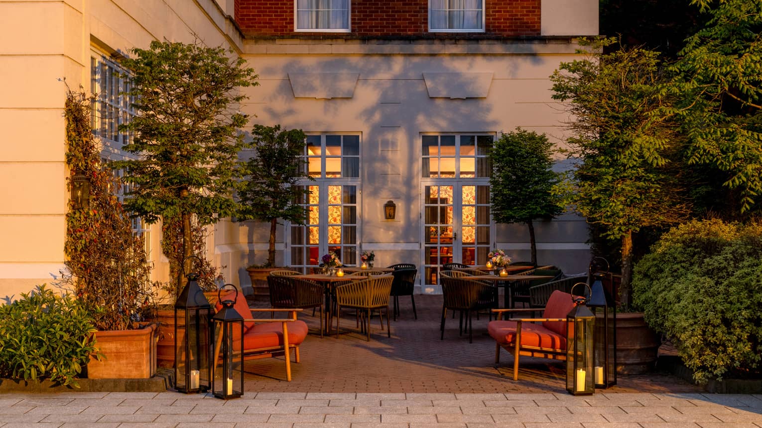 An outdoor dining space next to a building. Chairs and dining tables sit on a paved surface accented by trees and plantings.