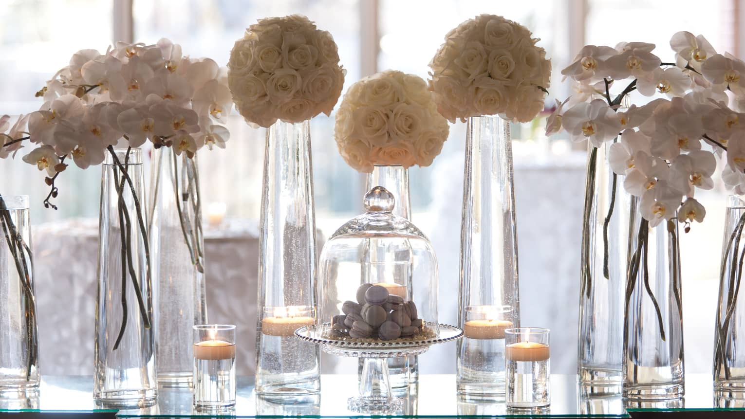 Flowers in tall glass vases, macarons in glass dessert display on table by window