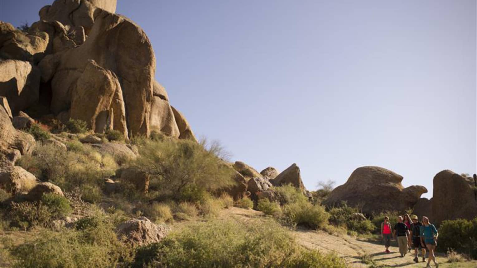 Hikers explore rugged mountain terrain, surrounded by desert flora and massive red sandstone and grey limestone rocks.