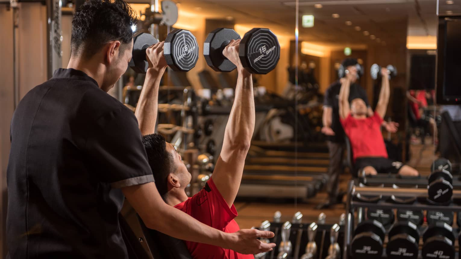 Man in chair in front of mirror lifts two 15 lb. weights above his head while personal trainer assists
